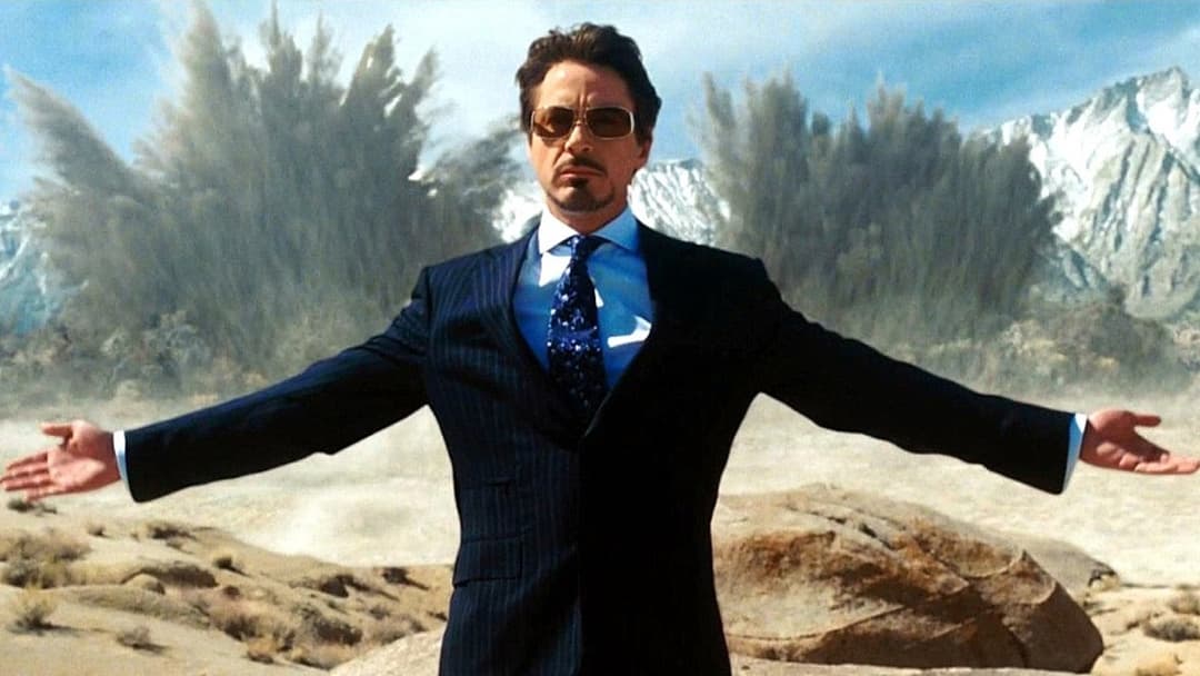 Robert Downey Jr en costume bleu ciel et veste noire, les bras écartés, face à des montagnes enneigées et des forêts de conifères