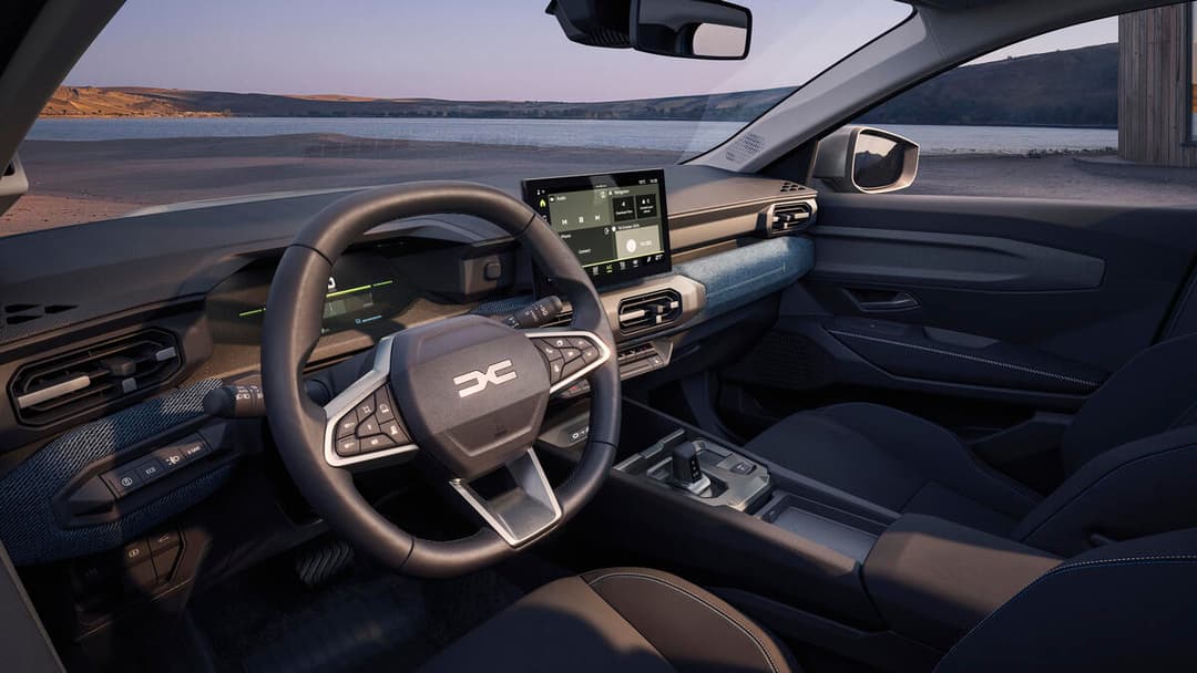 Intérieur du Dacia Striker avec écran tactile, volant noir, planche de bord grise et bleue, aérateurs horizontaux, vue sur lac en arrière-plan