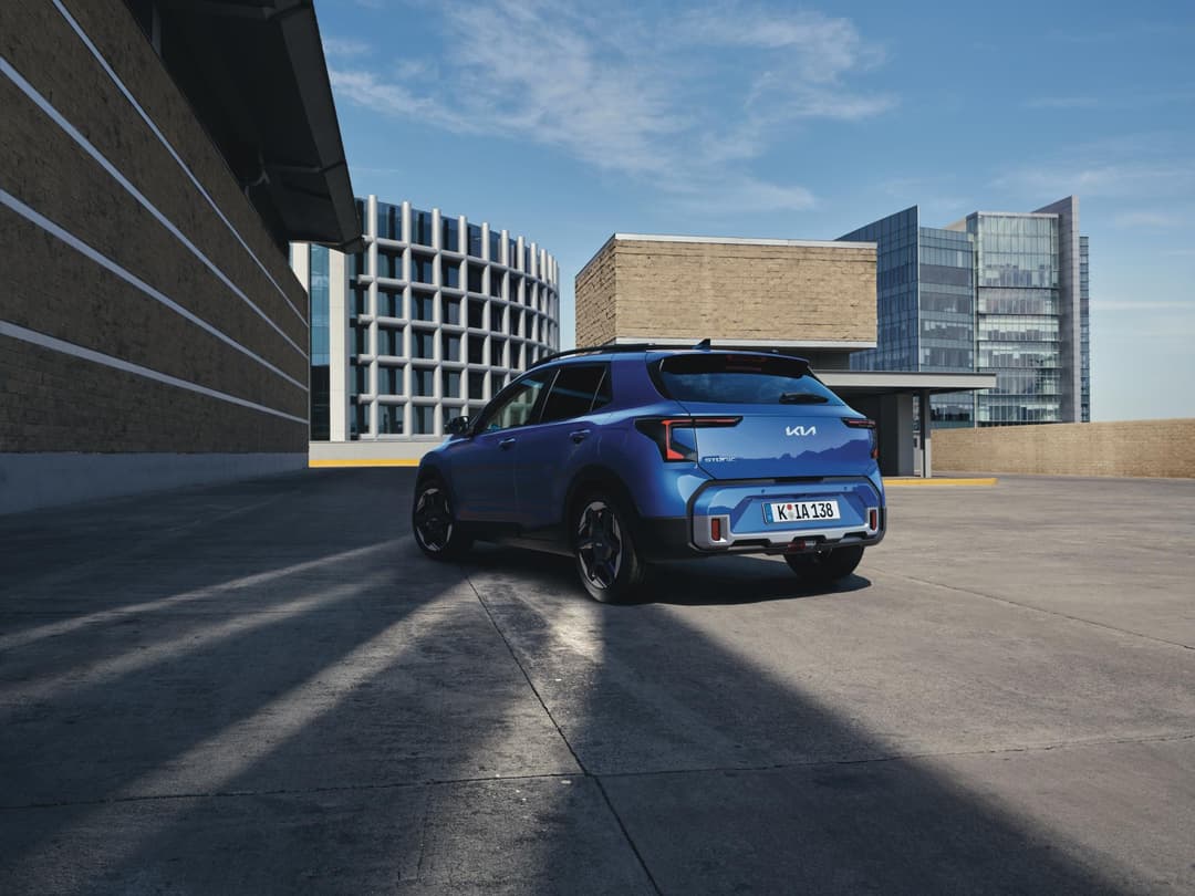 Kia Picanto bleu électrique vue de trois-quarts arrière, stationnée sur un parking urbain avec bâtiments modernes en arrière-plan