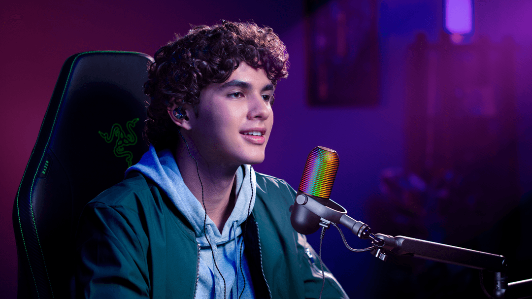 Streamer avec microphone Razer Seiren V3 Chroma aux effets RGB multicolores, assis dans un fauteuil gaming dans un décor aux lumières violettes