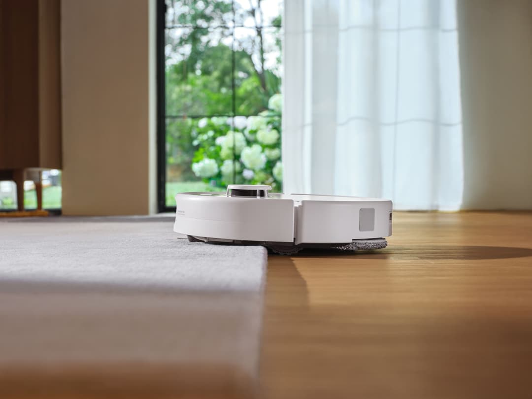 Roborock Qrevo 2 ProX blanc naviguant entre un tapis gris et un parquet clair, près d'une baie vitrée