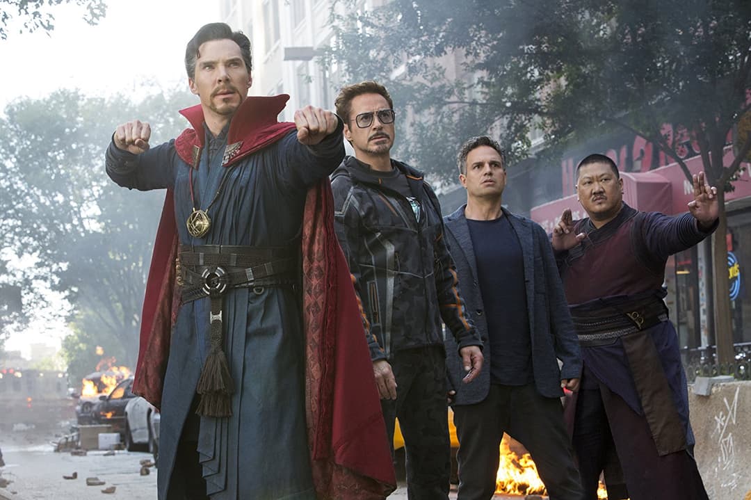 Cinq Avengers en costumes de combat debout dans une rue dévastée avec des explosions en arrière-plan