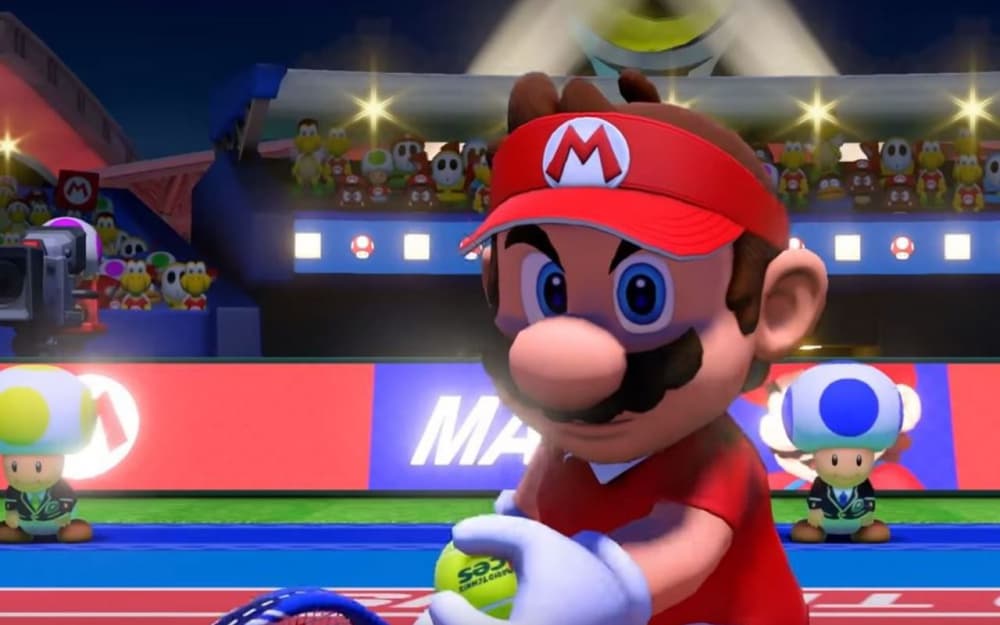 Mario en tenue rouge et casquette rouge tient une raquette et une balle de tennis jaune sur un court bleu et rouge, avec des spectateurs et des champignons en arrière-plan