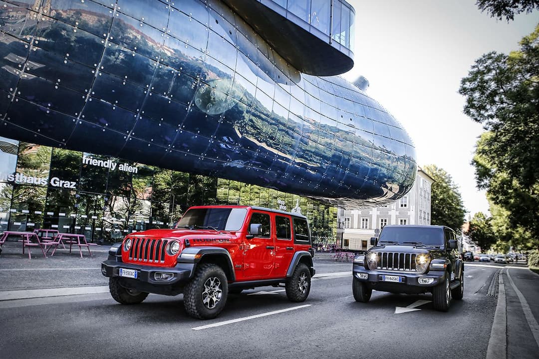 Deux Jeep Wrangler, l'une rouge et l'autre grise, garées sur une place urbaine devant un bâtiment moderne aux façades métalliques