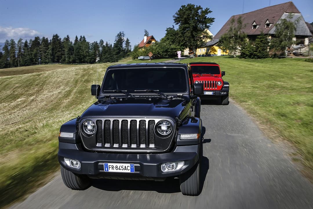 Jeep Wrangler bleu foncé en tête de convoi sur route de campagne, suivie d'une Jeep rouge, paysage alpin en arrière-plan