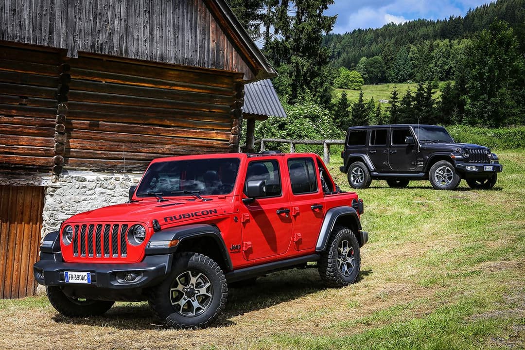 Jeep Wrangler rouge version Rubicon stationnée devant un chalet en montagne, avec un autre modèle noir en arrière-plan