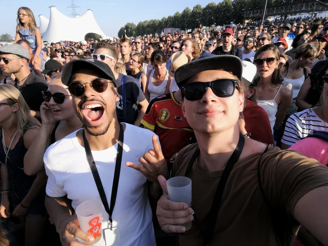 Deux hommes au premier plan sourient à la caméra au Paléo Festival Nyon, entourés d'une foule de festivaliers sous un ciel dégagé