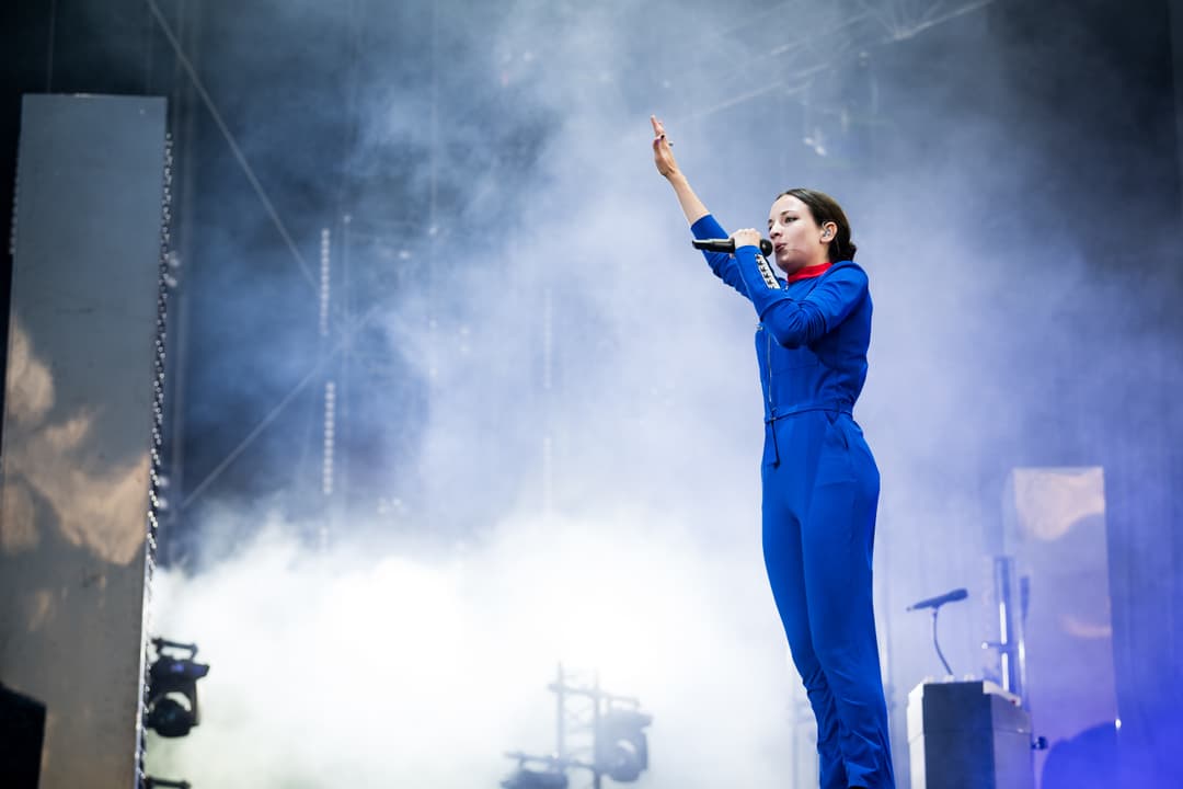 Jain en combinaison bleue sur scène au Paléo Festival, levant le bras vers le public dans une atmosphère enfumée