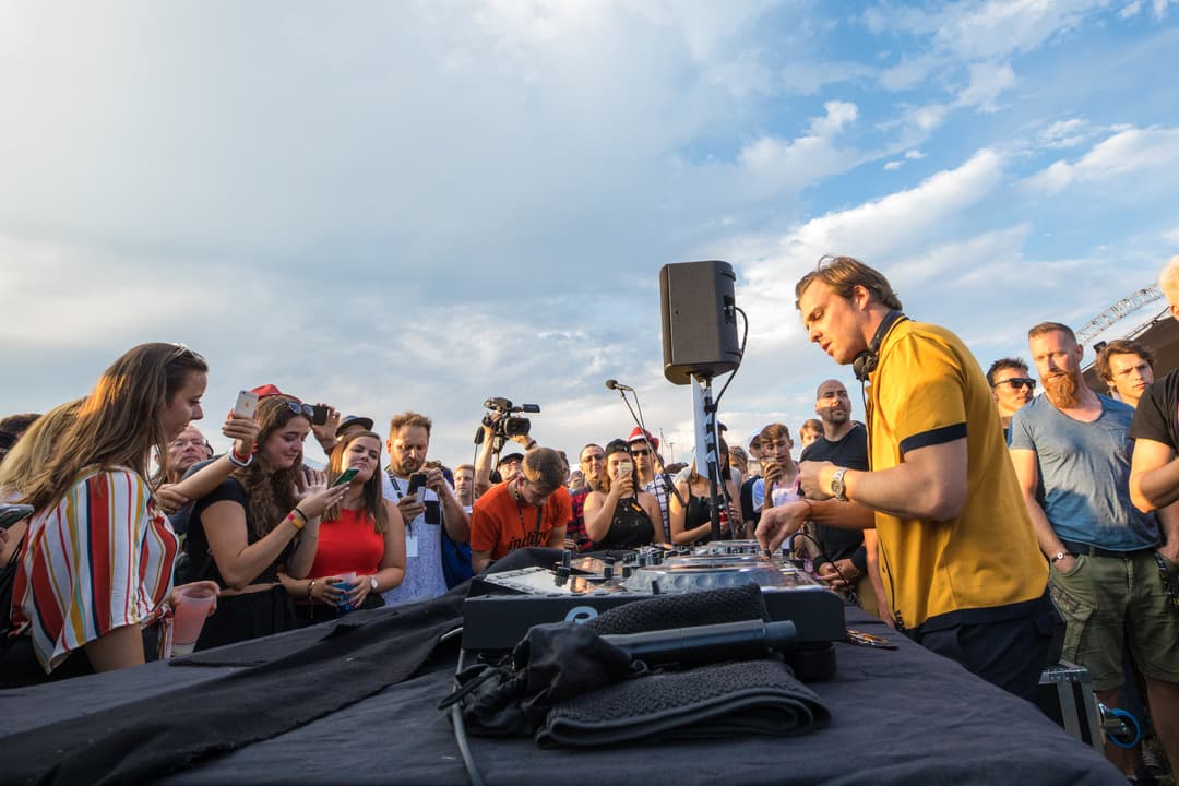 DJ en chemise jaune derrière les platines au Paléo Festival, entouré de spectateurs avec téléphones et appareils photo au coucher de soleil