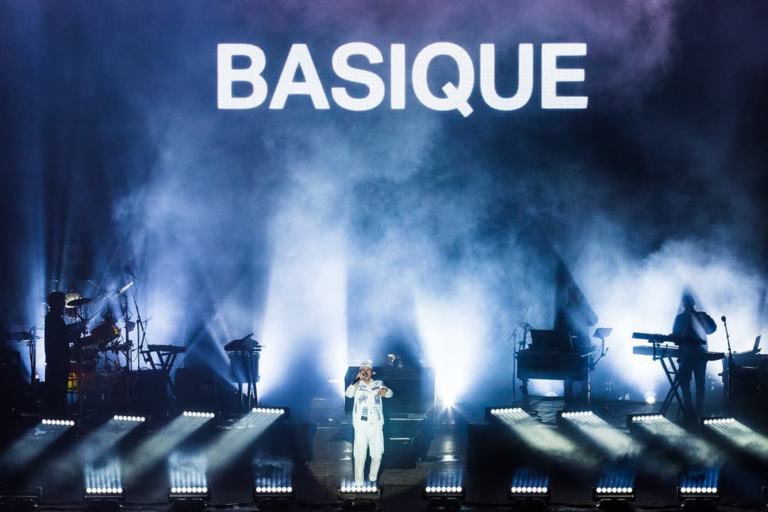 Basique sur scène au Paléo Festival, vêtu de blanc, entouré de musiciens et de jeux de lumière bleus intenses