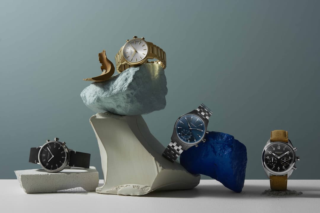 Quatre montres Kronaby Sweden avec bracelet rigide, présentées sur des formes géométriques en pierre et tissu, fond gris-bleu