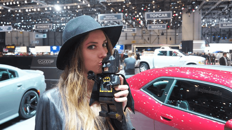 Femme tenant une figurine Batman au Salon de l'Automobile de Genève, avec des voitures rouges et blanches en arrière-plan