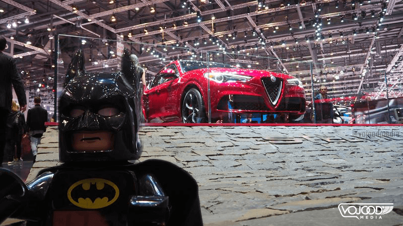 Batman en costume noir face à une Alfa Romeo rouge au Salon International de l'Automobile de Genève, sous les projecteurs du hall d'exposition