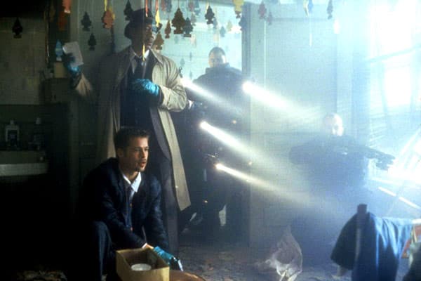 Détectives inspectant une scène de crime dans un décor sombre et poussiéreux, éclairé par des rayons de lumière blanche
