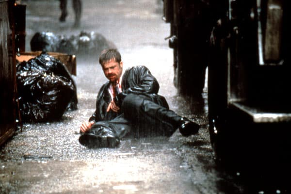 Un homme assis sous la pluie dans une ruelle sombre, entouré de sacs-poubelle noirs, ambiance sinistre et urbaine