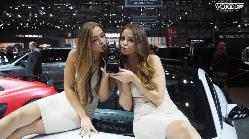 Deux hôtesses présentent le logo Batman sur un téléphone au Salon de l'Automobile de Genève, assises sur une voiture rouge