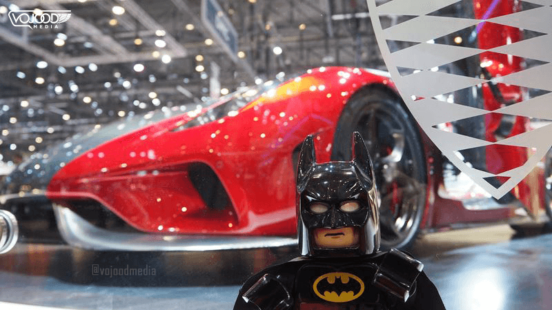 Figurine Lego Batman en premier plan devant une voiture de sport rouge brillante dans un hall d'exposition