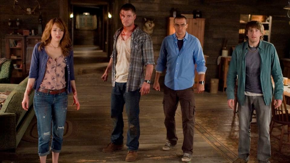 Cinq jeunes adultes traversent l'intérieur d'une cabane en bois, vêtus de façons différentes suggérant des archétypes de film d'horreur