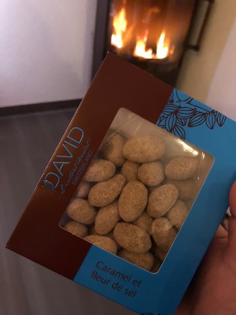 Boîte David l'Instant Chocolat contenant des amandes enrobées de chocolat au caramel et fleur de sel, tenue en main devant une cheminée
