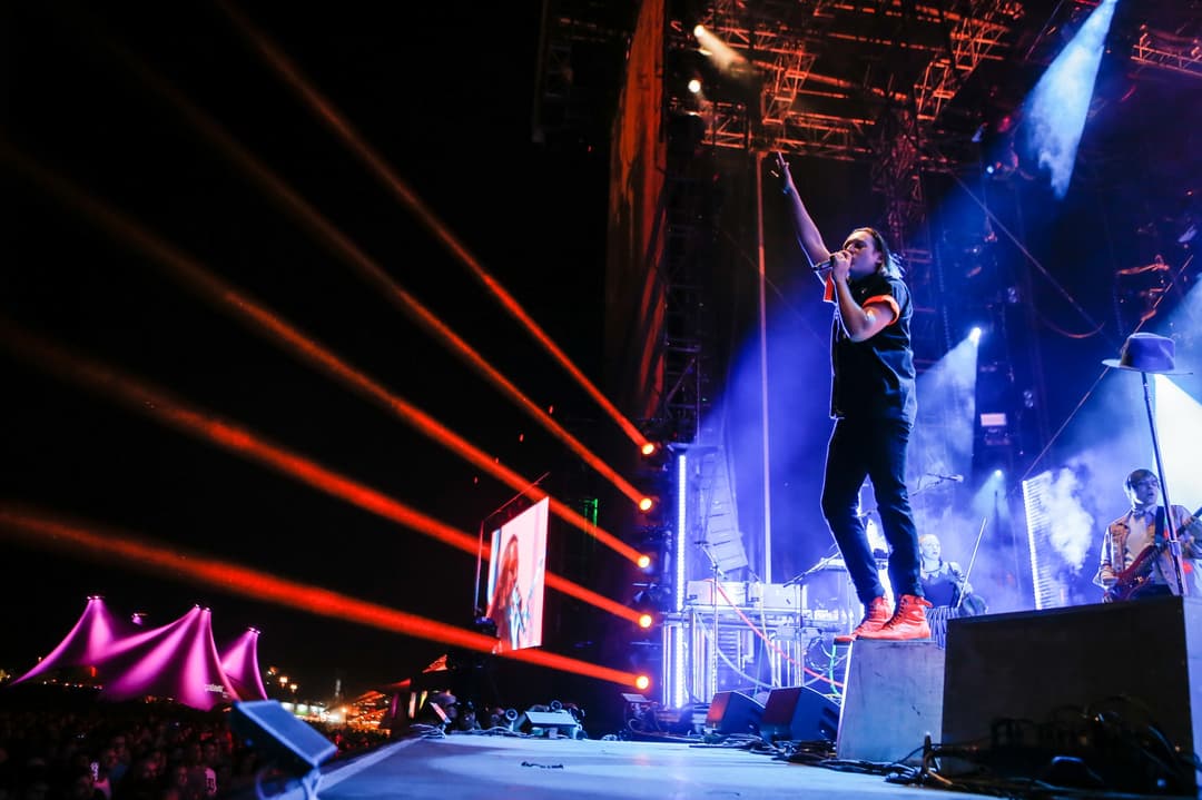 Chanteur d'Arcade Fire sur scène au Paléo Festival, éclairages bleus et rouges, musiciens visibles en arrière-plan