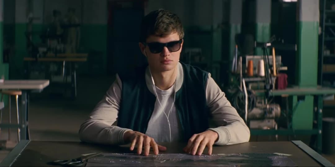 Baby, jeune braqueur de banque portant des lunettes de soleil noires, assis à une table dans un garage avec les mains posées devant lui