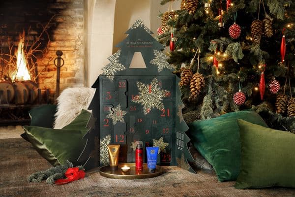 Calendrier de l'Avent Rituals de forme maison verte décorée de flocons d'or, entouré de sapins, bougies et coussins verts dans un décor hivernal