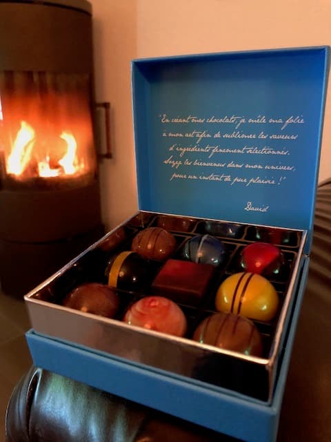 Écrin bleu ouvert contenant des chocolats colorés artisanaux de David l'Instant Chocolat, présentés avec une cheminée allumée en arrière-plan