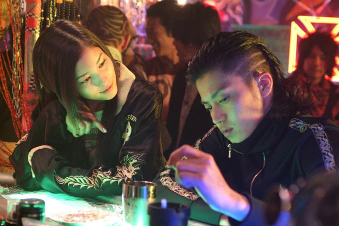 Deux jeunes délinquants regardent des tatouages sous lumière verte et rose dans un bar de Crows Zero