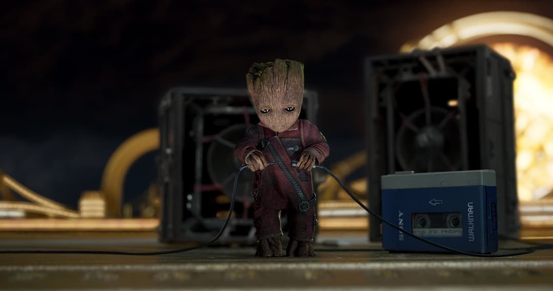 Groot, petit personnage arborescent aux yeux lumineux vêtu de rouge, tient un lecteur cassette bleu vintage parmi des équipements technologiques
