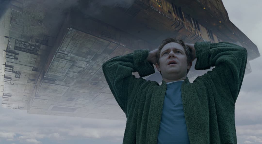 Martin Freeman en costume vert kaki regarde vers le ciel, une immense structure spatiale planant au-dessus de lui