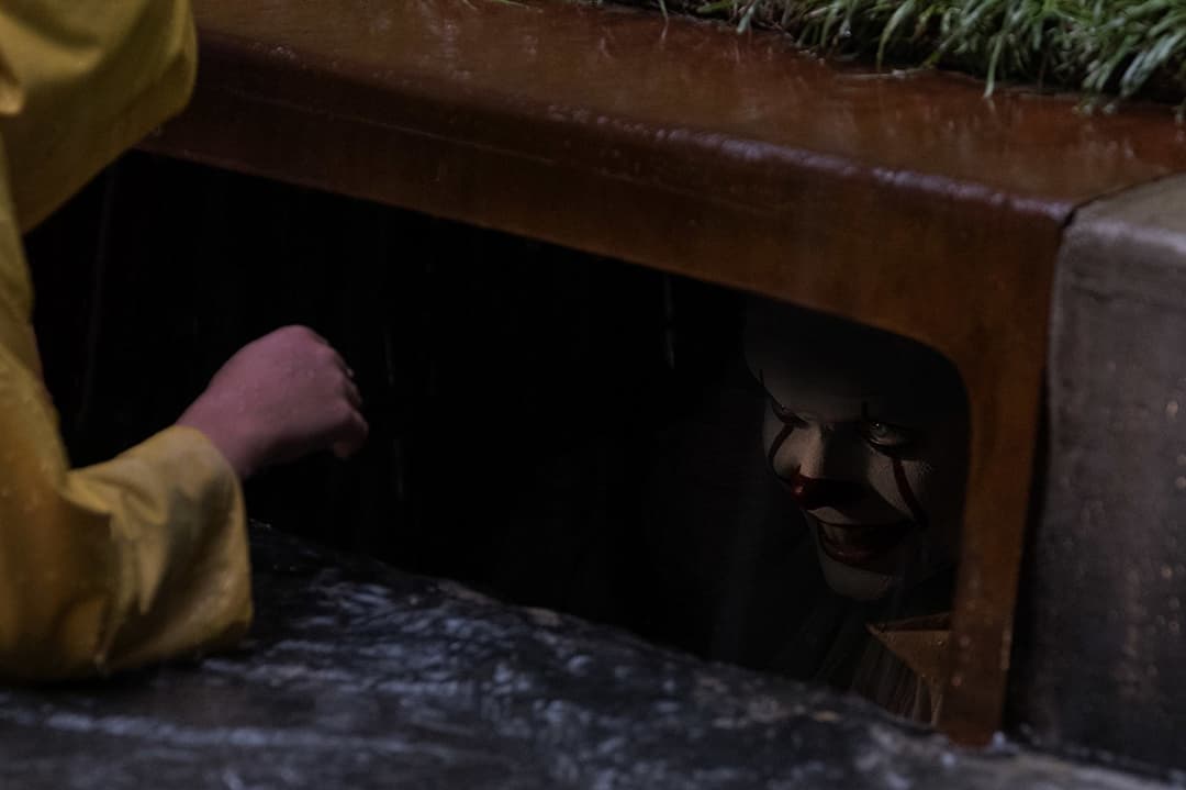 Un enfant en imperméable jaune se penche vers une bouche d'égout où apparaît un visage grimaçant dans l'obscurité