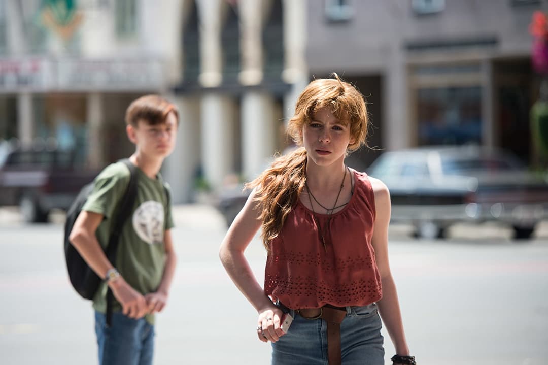 Beverly, personnage du film Ça, vêtue d'un chemisier rouge, marche dans une rue ensoleillée tandis qu'un garçon la suit