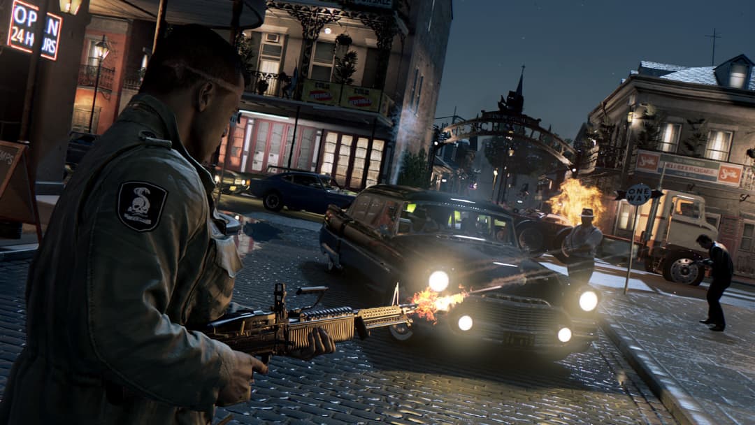 Personnage tirant à la mitrailleuse dans une rue urbaine la nuit, avec explosions et véhicules en arrière-plan dans Mafia III