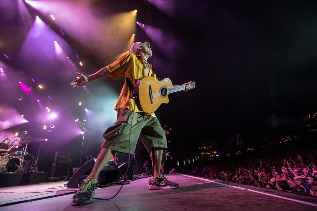 Manu Chao en concert au Paléo Festival 2017, guitare à la main, sous des projecteurs pourpres et jaunes, face à une foule nombreuse