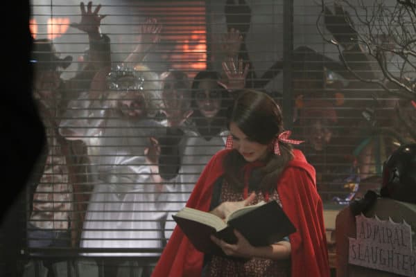 Femme en manteau rouge lisant un livre devant une fenêtre avec des zombies qui pressent leurs mains contre le verre