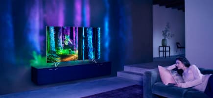 Téléviseur Philips AmbiLux 4K avec Ambilight projetant un halo coloré bleu, vert et violet sur le mur, dans un salon avec canapé
