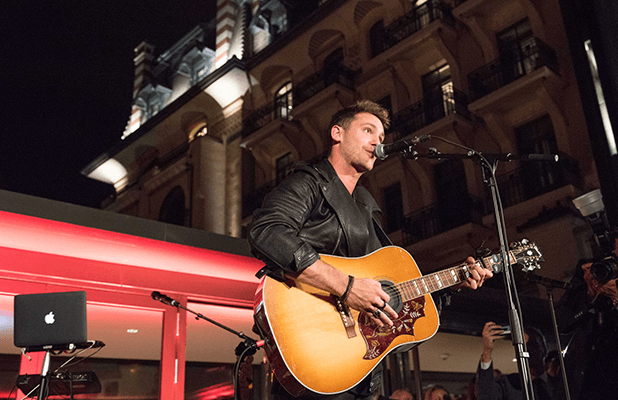 Bastian Baker chanteur jouant de la guitare acoustique sur scène, façade illuminée de l'Hôtel Royal Savoy Lausanne en arrière-plan