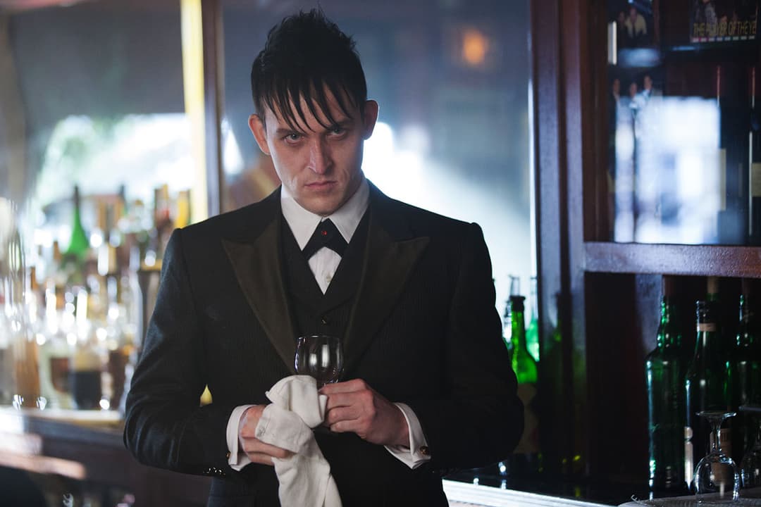 Le Pingouin en costume noir et nœud papillon, tenant un verre dans un bar sombre rempli de bouteilles