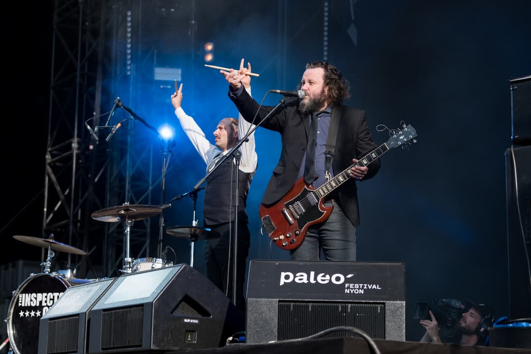 Musiciens sur scène au Paléo Festival Nyon, l'un jouant de la guitare, l'autre au micro, éclairage bleu intense en arrière-plan